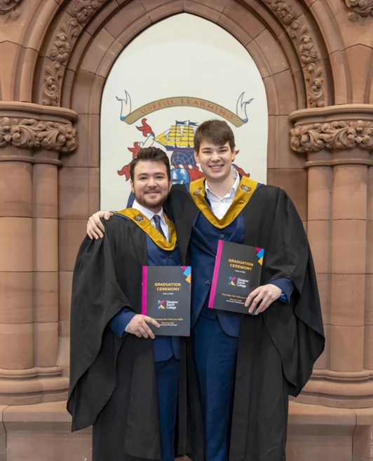 Two graduates smiling and holding certificates in front of the Glasgow Kelvin College graduation crest backdrop Two graduates smiling and holding certificates in front of the Glasgow Kelvin College graduation crest backdrop